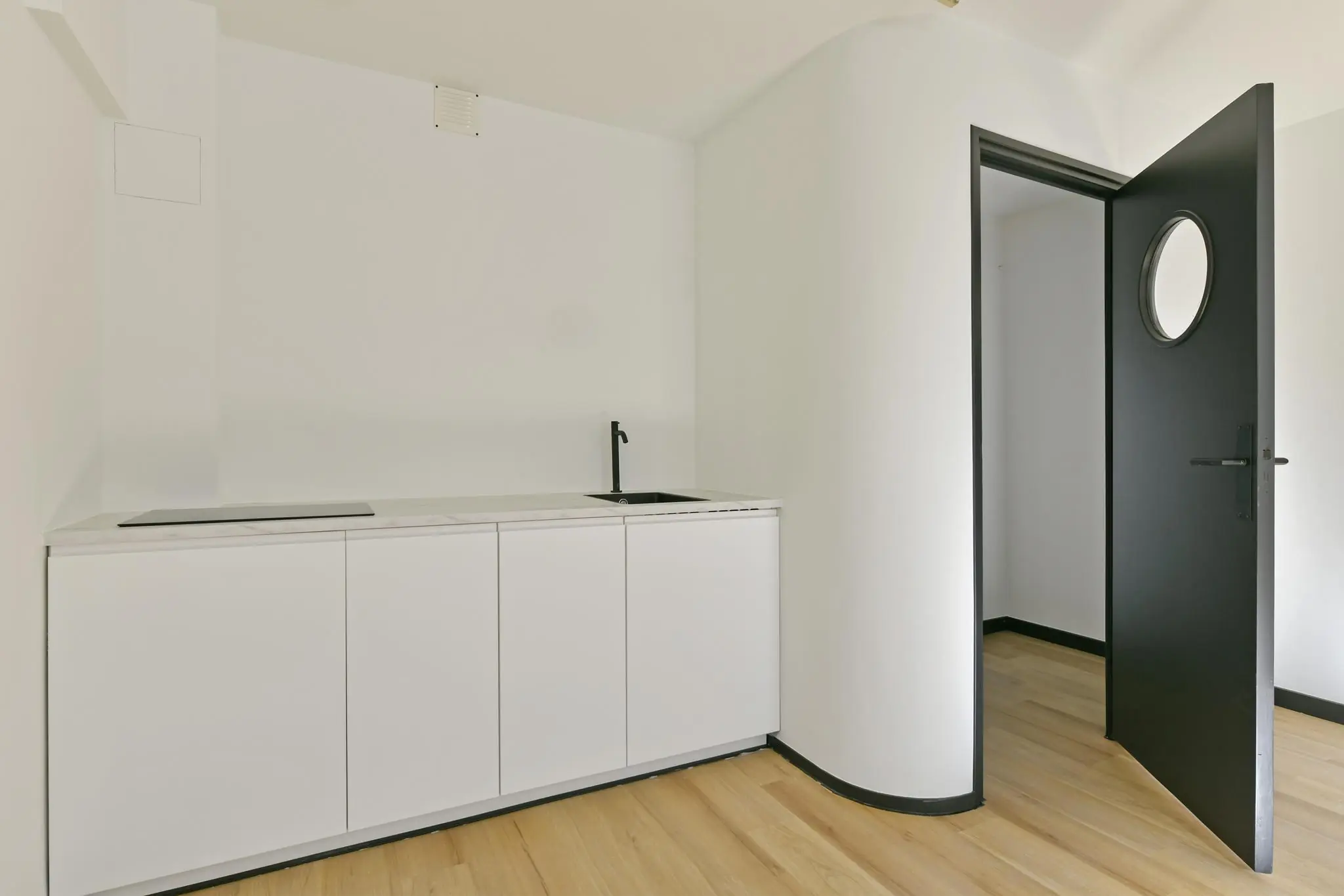 Minimalistische kitchenette aan de Cornelis Dopperkade met witte kasten, zwarte kraan en open zwarte deur met rond raam.